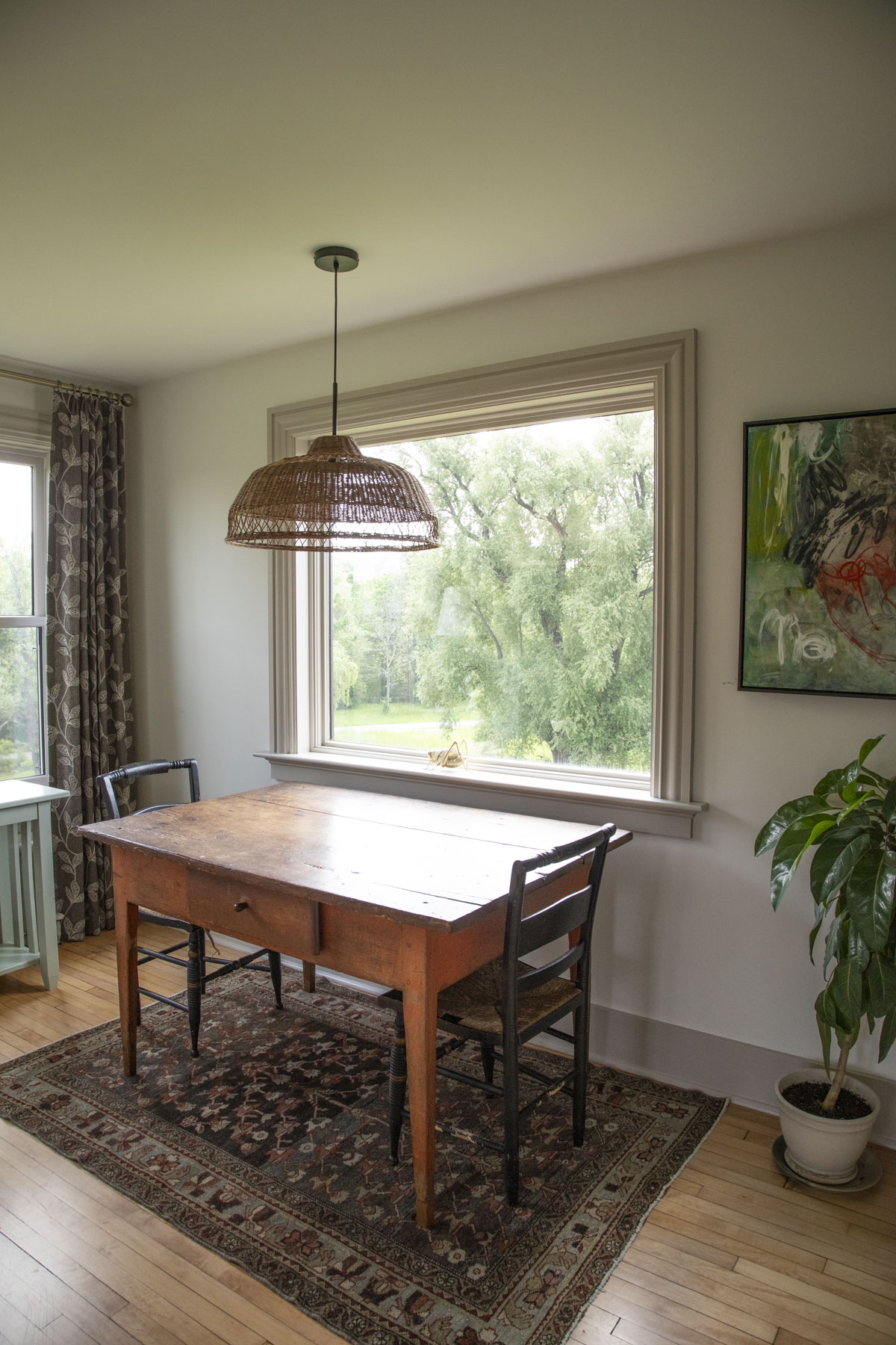 an antique table under a picture window with a large wicker light fixture hung over it. 